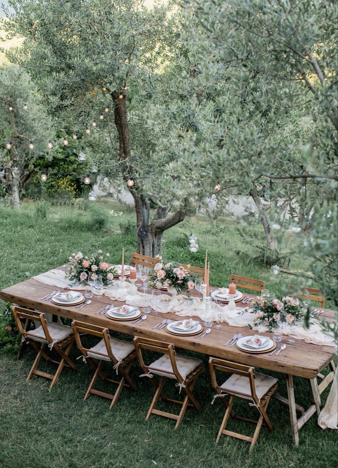 Long wooden table set for dinner under string-lit olive trees, with blush roses and taper candles