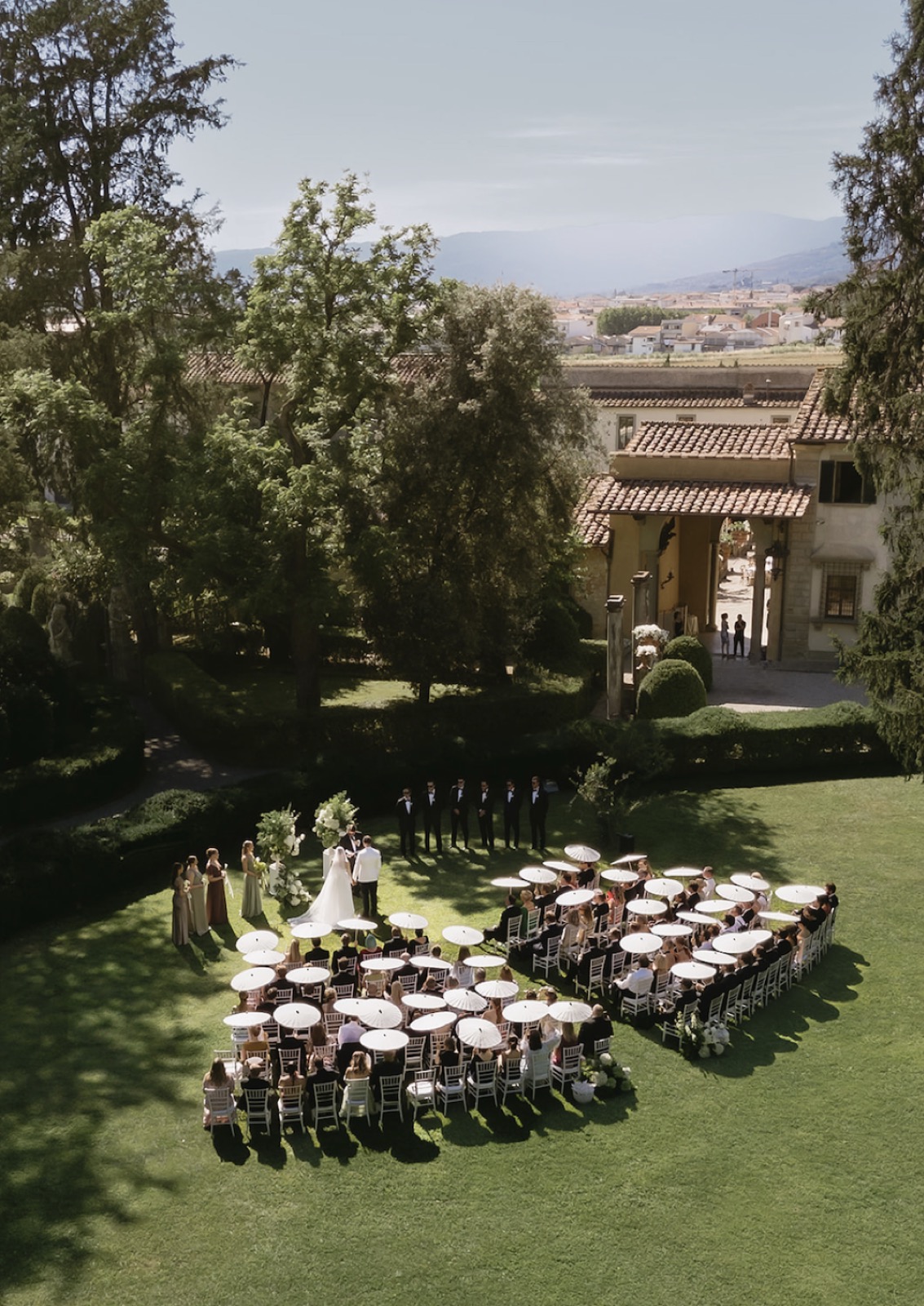 Aerial view of a wedding ceremony with guests seated under white parasols in a villa garden