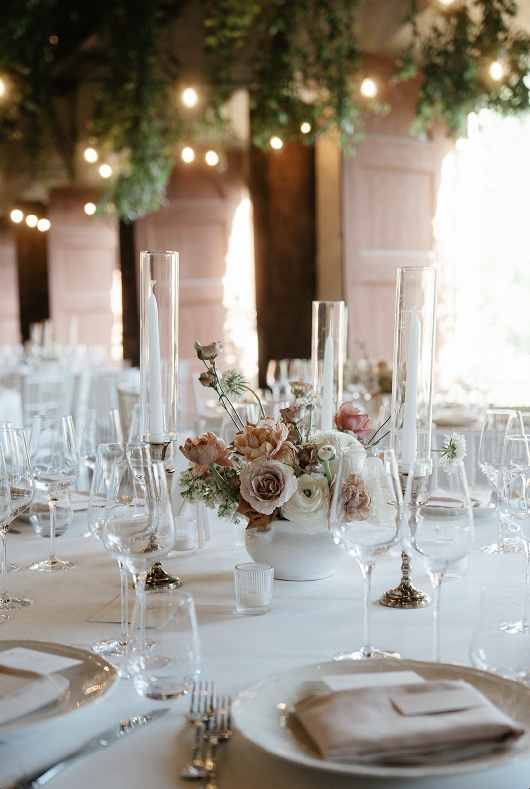 Reception dinner table with dusty rose florals, glass taper candle holders, and string lights under a loggia