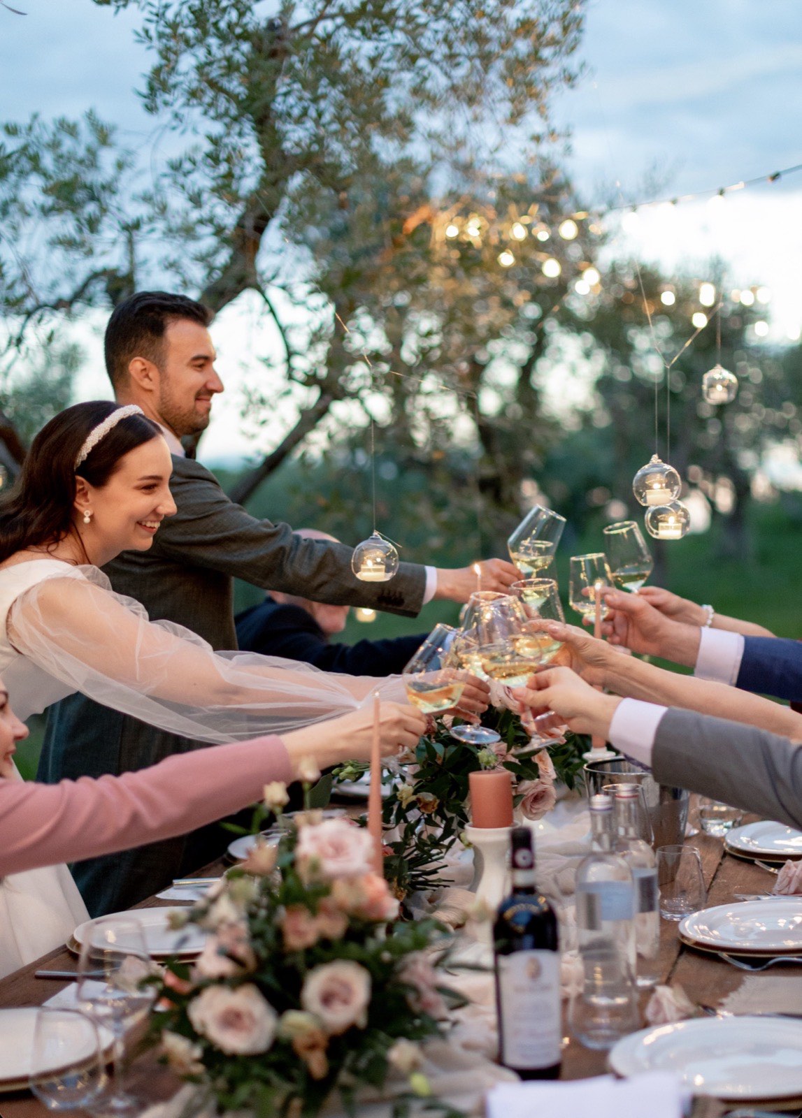 Wedding guests raising a toast at an olive-grove dinner table lit with string lights and hanging candles
