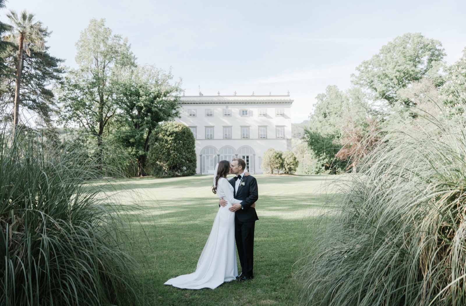 Bride and groom kissing on the lawn in front of a grand villa, framed by ornamental grasses